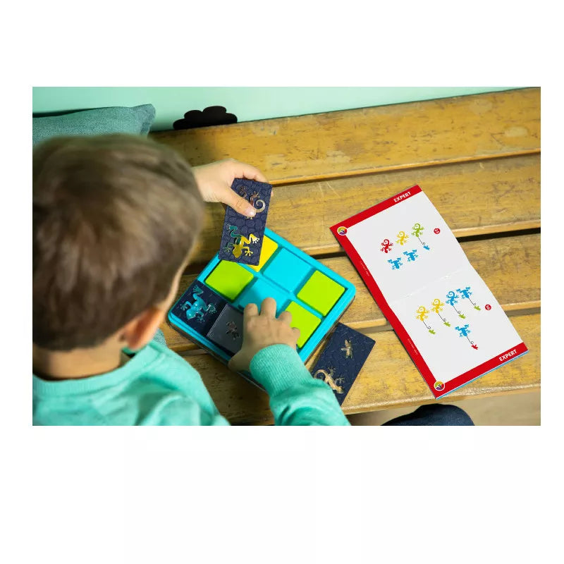 A child in a green sweater is deeply engaged with the SmartGames Colour Catch DAMAGED BOX, an educational puzzle game that helps improve problem-solving skills through play. They are fitting colorful pieces into a grid while using the illustrated solution book for guidance on the wooden surface beside them.