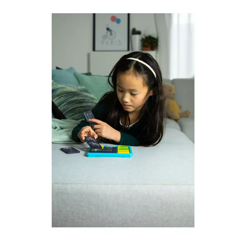 A young girl with long dark hair and a headband lounges on a sofa, focused on solving the SmartGames Colour Catch DAMAGED BOX puzzle on her vibrant, rectangular electronic device. A teddy bear can be seen in the background. The room exudes coziness, with its soft color palette enhancing the peaceful atmosphere.