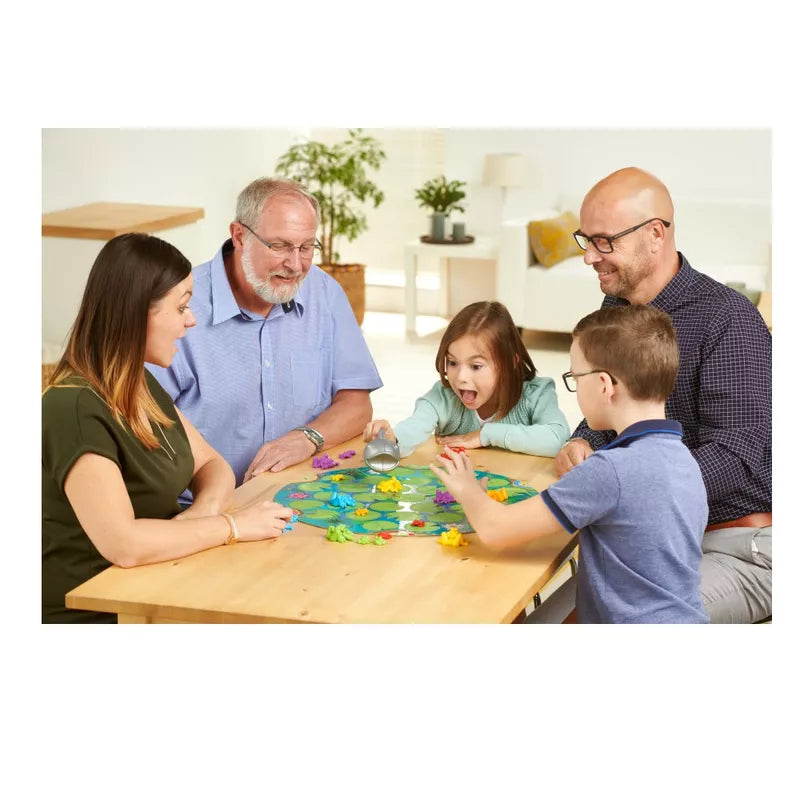 A frog family of five, including two men, a woman, and two children, is engaged and smiling while playing SmartGames Froggit around a table. The room is well-lit with lush plants and cozy furniture in the background, showcasing clever planning and flexible thinking.