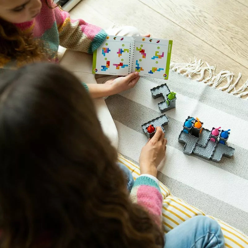 Two people sit cross-legged, deeply focused on SmartGames Tower Stacks. A booklet with colorful patterns guides them as puzzle pieces spread over a checkered mat with vibrant figures. Both wear bright sweaters, matching the playful problem-solving session perfectly.