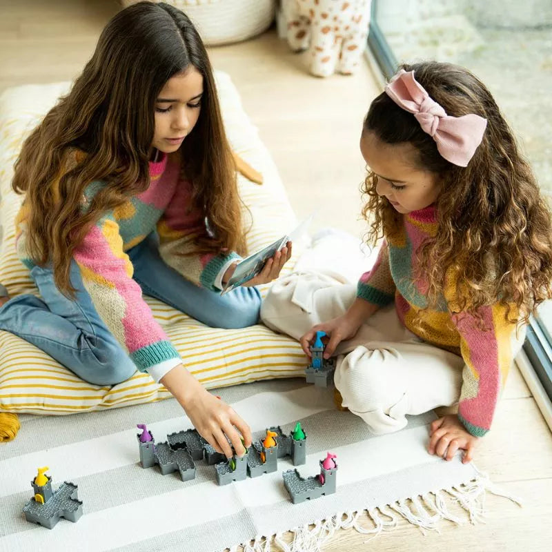 Two children with long hair play SmartGames Tower Stacks on a striped rug by the window. Both wear colorful sweaters. One child holds a game piece while the other is engrossed in problem-solving on a phone. A pillow and patterned blanket add coziness to the scene.