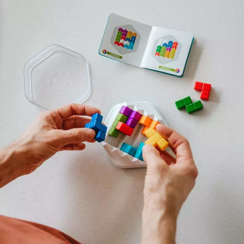 Hands expertly assemble colorful pieces from the SmartGames Zig Zag Puzzler on a white table, guided by an open instruction booklet with completed examples. Nearby lies a clear container lid and unused pieces, sparking creativity and problem-solving skills.