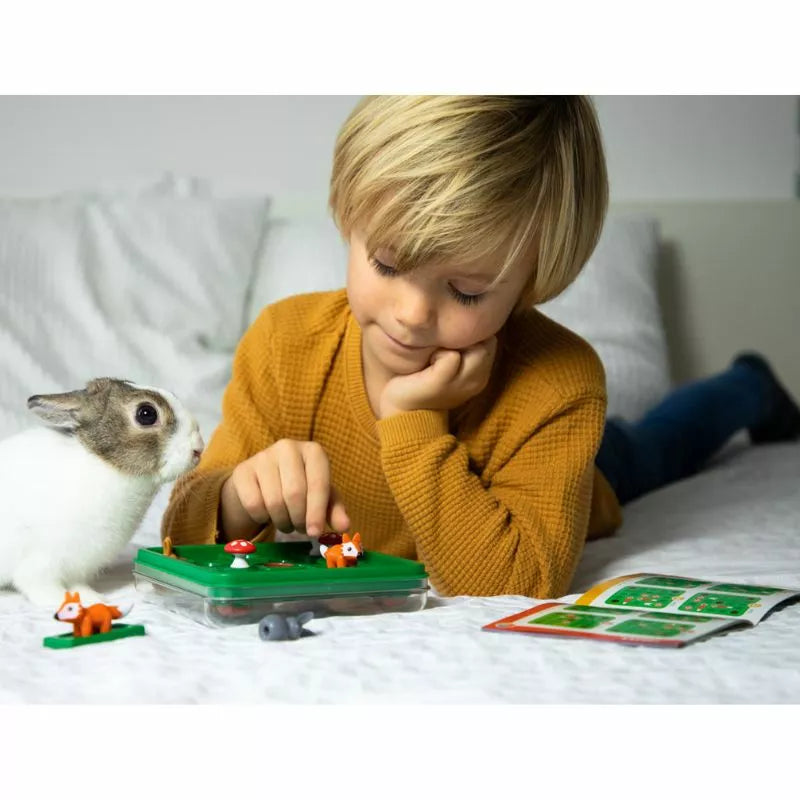 A young child with blonde hair, dressed in a mustard-colored sweater, is lying on a bed absorbed in the Smartgames Jump In' green puzzle game, which boosts spatial insight. Next to them is a small white and gray rabbit, while an illustrated booklet resembling a delightful travel game moment remains open nearby.