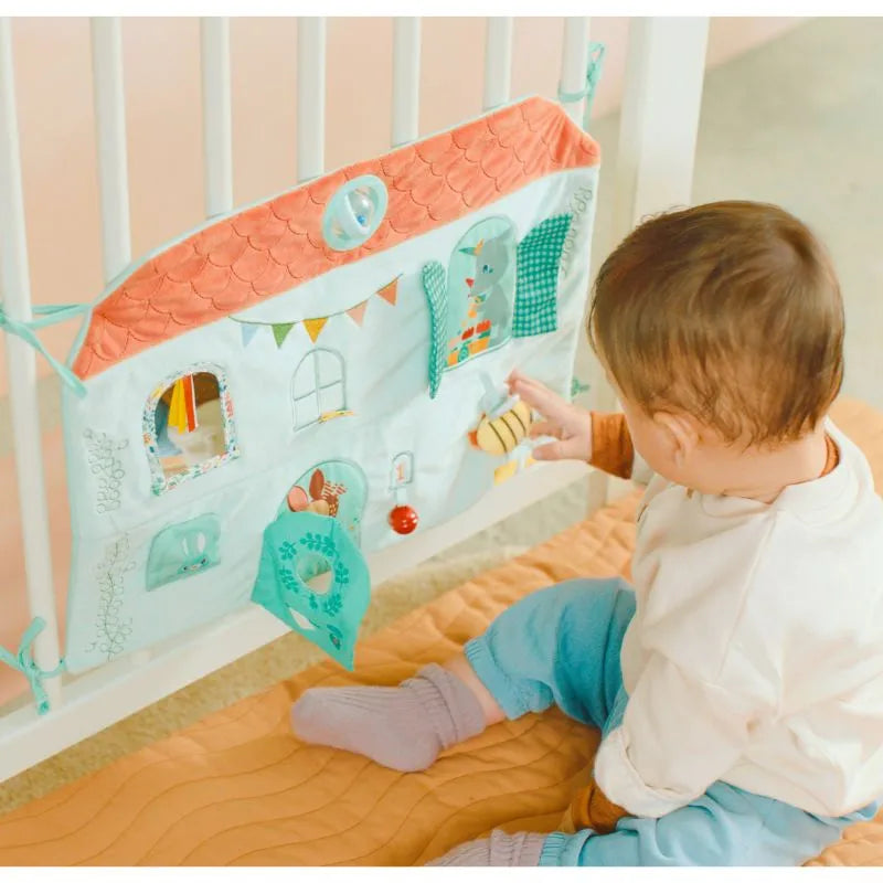 A baby sits on an orange mat enjoying the Lilliputiens I'm Discovering Stella & Joe House Soft Discovery Panel, a perfect first toy or gift. Designed like a house, it features various textures and interactive elements like flaps, buttons, and a detachable toy.