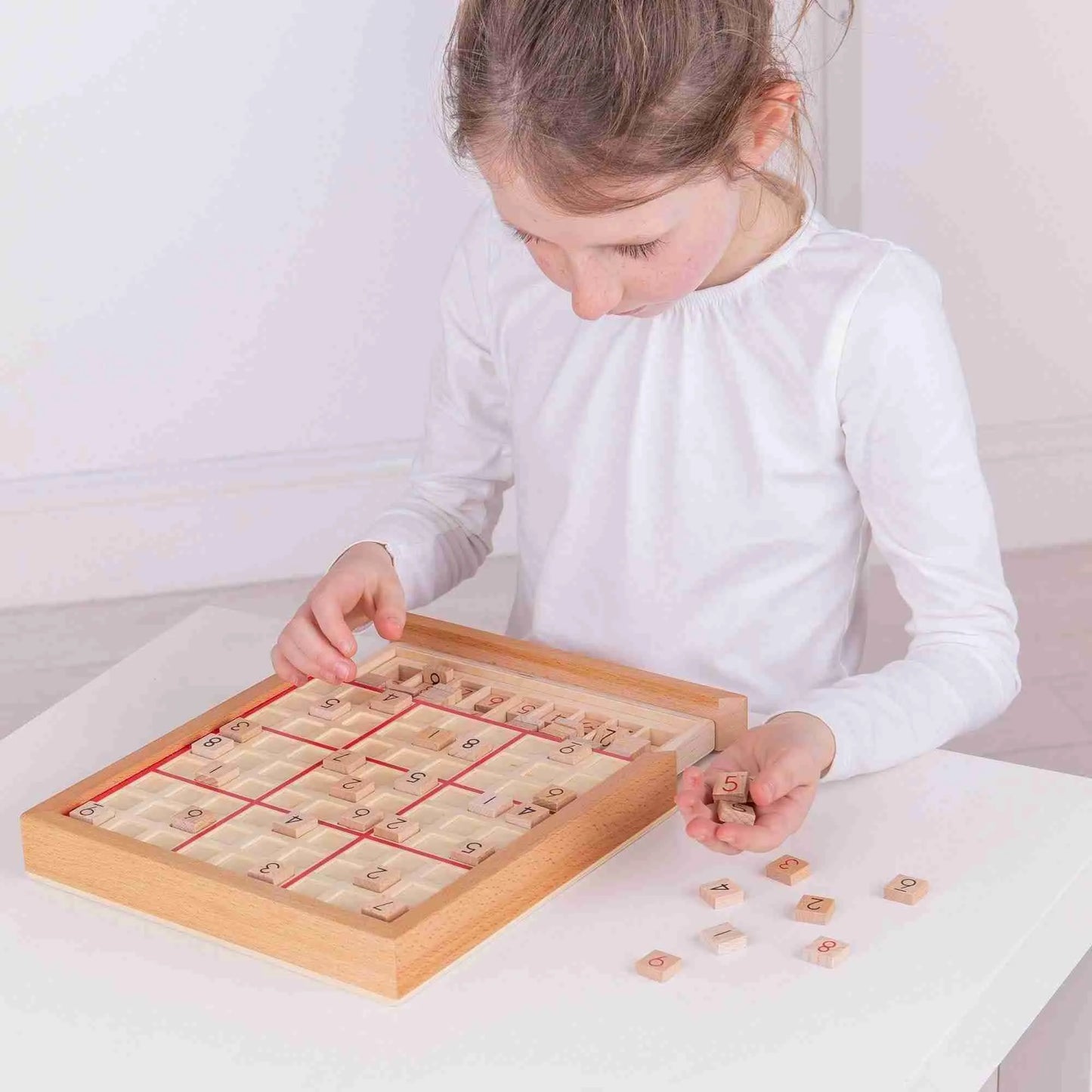 A child in a white long-sleeve shirt is deeply engaged in a strategic game, playing with the Bigjigs Sudoku Game on the table. This brain-training activity features numbered tiles arranged in a grid, with several left outside, providing both fun and concentration.