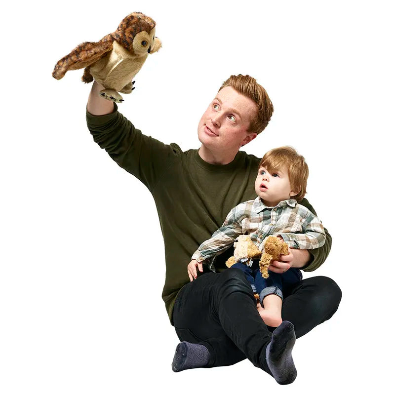 A reddish-brown-haired man in a green sweater and black pants sits cross-legged with a toddler in a plaid shirt. The child holds a stuffed animal as the man animates "The Puppet Company Hide-Away Tawny Owl" glove puppet, both gazing at it with delight.