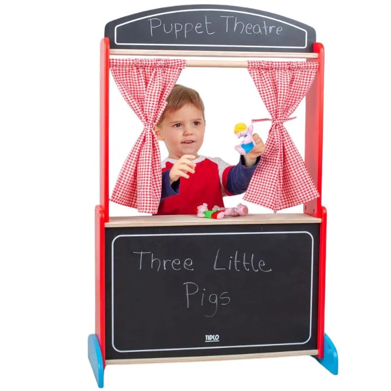 A child stands behind the vibrant Tidlo Wooden Puppet Theatre with red checkered curtains, holding a puppet in each hand. The chalkboard at the bottom reads "Three Little Pigs," setting the stage for imaginative play. The frame of the theater is painted in lively red and blue hues.