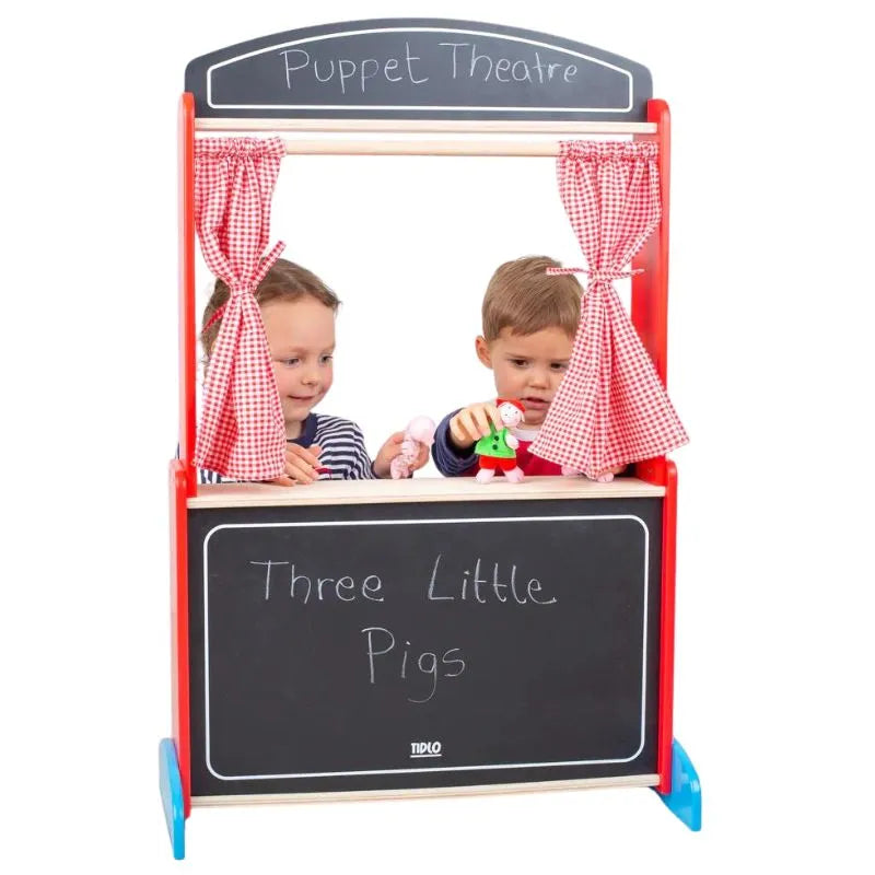 Two children are engaged in imaginative play with puppets on a Tidlo Wooden Puppet Theatre. A chalkboard reads "Three Little Pigs." The children hold pig and wolf puppets, performing a lively show. Red gingham curtains frame the theater stage, adding charm to their performance.