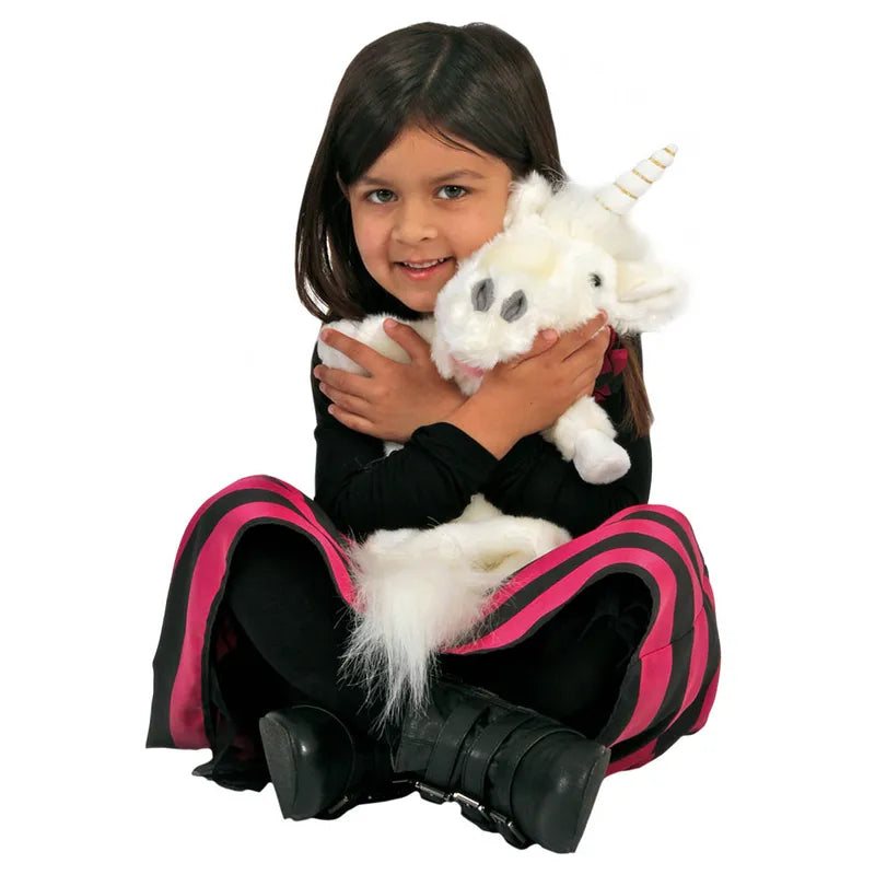 A young girl with long dark hair, dressed in a black outfit with pink stripes and black boots, sits cross-legged while hugging The Puppet Company Long Sleeved Puppet Unicorn—a plush white unicorn hand puppet with a yellow horn. The girl smiles at the camera.