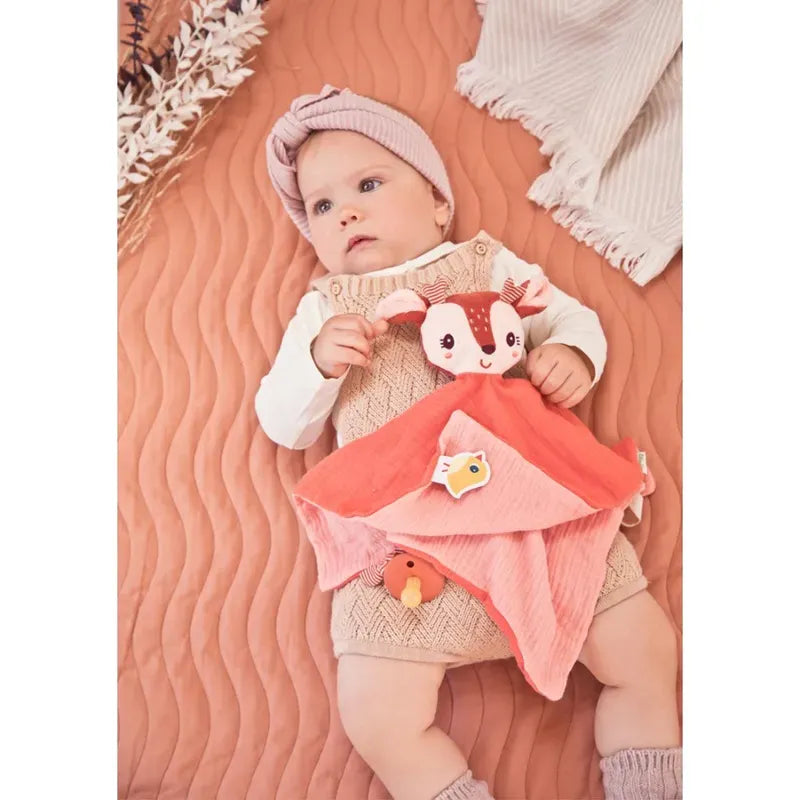 A baby lies on a quilted peach-colored surface, wearing a beige knitted romper over a white long-sleeve shirt and a pink headwrap. The baby holds Lilliputiens Stella the Fawn Comforter. Nearby is an organic comforter with fringed edges and some dried flowers, adding to the cozy scene.