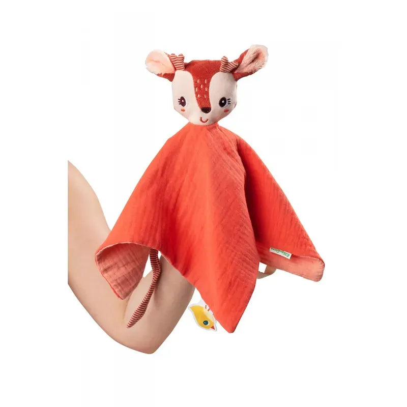 A person holding a red animal-themed Lilliputiens Stella the Fawn Comforter featuring a cute, smiling deer face with fabric ears and antlers. The Lilliputiens Stella the Fawn Comforter is draped over the person's arm and has a soft, textured fabric that's machine-washable for easy care.