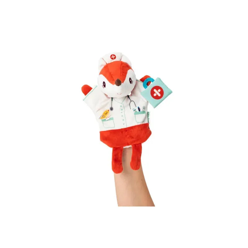 A person holds up a Lilliputiens Alice Doctor Handpuppet designed as a nurse fox, perfect for imaginative play. The fox wears a white lab coat with medical symbols and a stethoscope; its paws are adorned with red accents. One paw holds a small blue bag with a red cross, making it ideal for kids aged 2 years+.