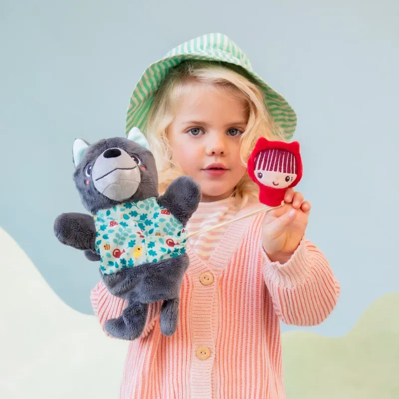 A young child with blonde hair, wearing a green-striped hat, pink cardigan, and striped shirt, holds up a Lilliputiens Louis Wolf Handpuppet in one hand and a Little Red Riding Hood Finger Puppet in the other. The background is softly colored in pastel shades, perfect for imaginative and creative play.