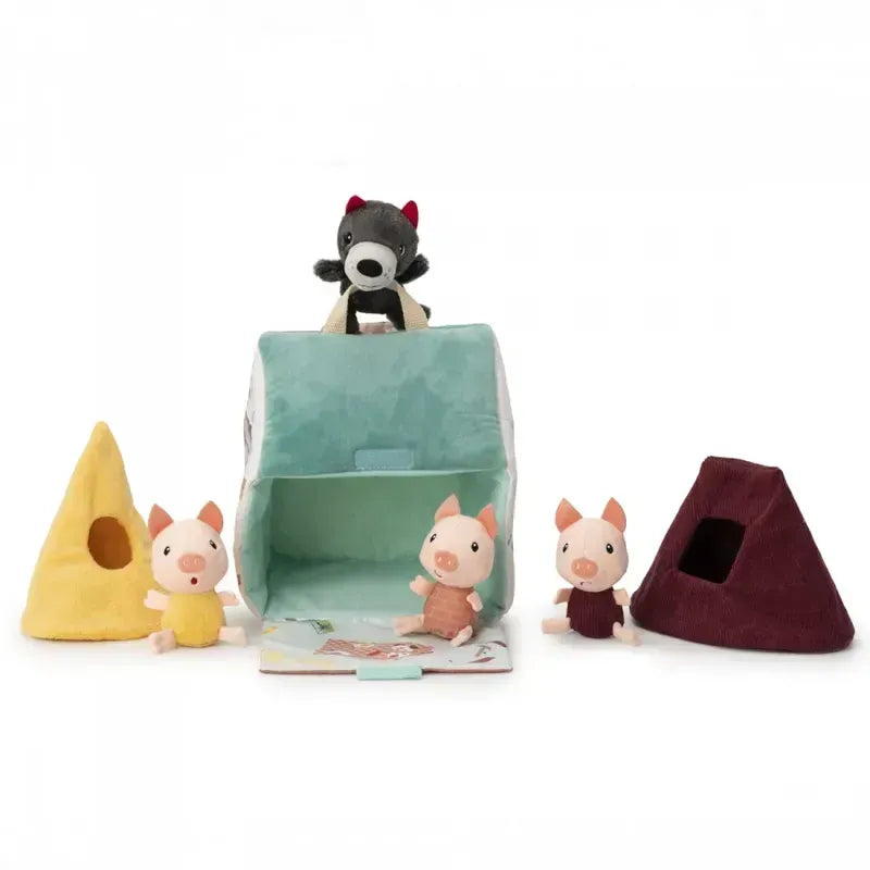 The Lilliputiens Houses Of The Wolf And The 3 Little Pigs fabric toy set includes a wolf perched on a turquoise hut, three pigs dressed in colorful outfits (yellow, orange, and brown), two pyramid-shaped houses (one yellow and one brown), and a small book to inspire creative play and enhance fine motor skills.