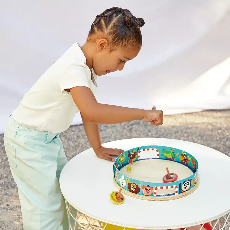 A child with braids and a white shirt leans over a round table, playing with the Janod Applepop - Spinning Top Arena inside a circular track decorated with animal illustrations. The table features a white surface and a metal wireframe base, enhancing the charm of this delightful children's toy.