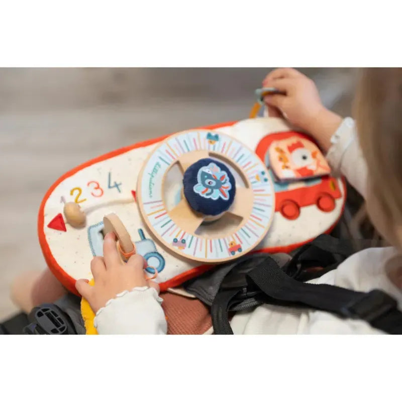 Sure, here is the sentence with the product name included.

A child holds a colorful Lilliputiens Farm Car Activity Panel. The baby toy features various elements, including a spinning wheel with a blue cat figure, numbers from 1 to 4, and a red car illustration. The child is dressed in white and focused on playing with the Lilliputiens Farm Car Activity Panel during their car journeys.