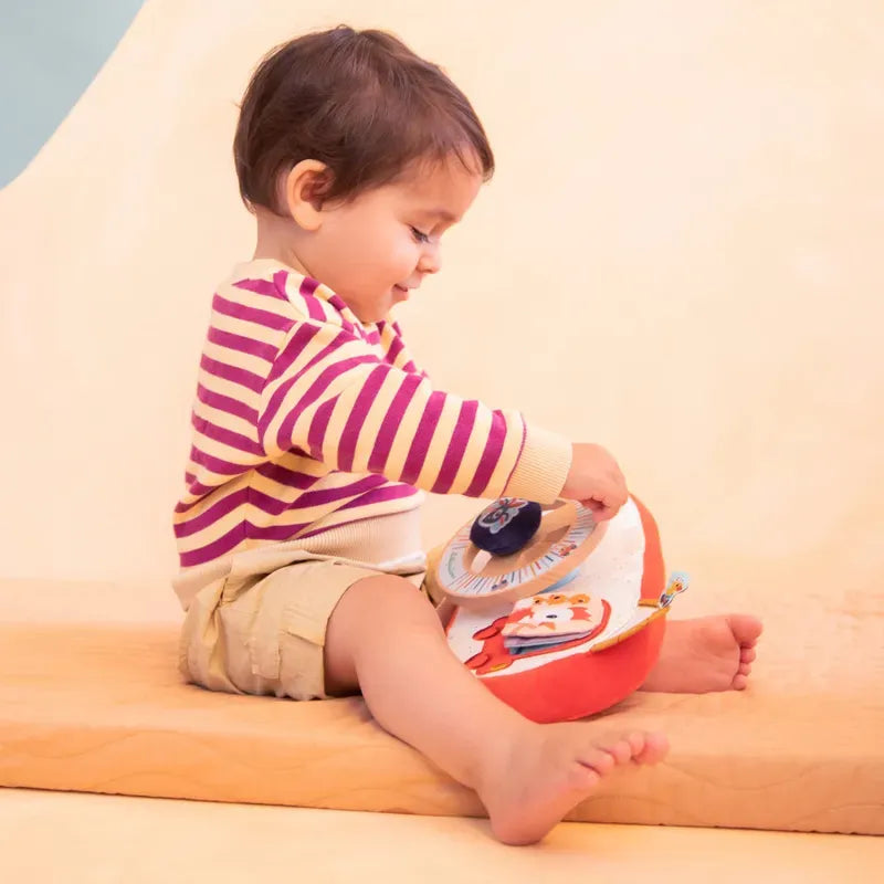 A toddler with short brown hair is seated on a cushioned surface, wearing a striped sweater and beige shorts. They are playing with a colorful Lilliputiens Farm Car Activity Panel, an educational baby toy featuring illustrations and interactive elements, perfect for car journeys.