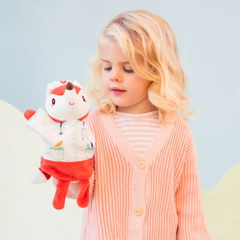 A young child with blonde hair, wearing a light pink cardigan, holds a Lilliputiens Alice Doctor Handpuppet. The puppet resembles a fox in a white lab coat and red pants, complete with a stethoscope accessory, perfect for imaginative play. Ideal for children aged 2 years+, it invites hours of creative storytelling.