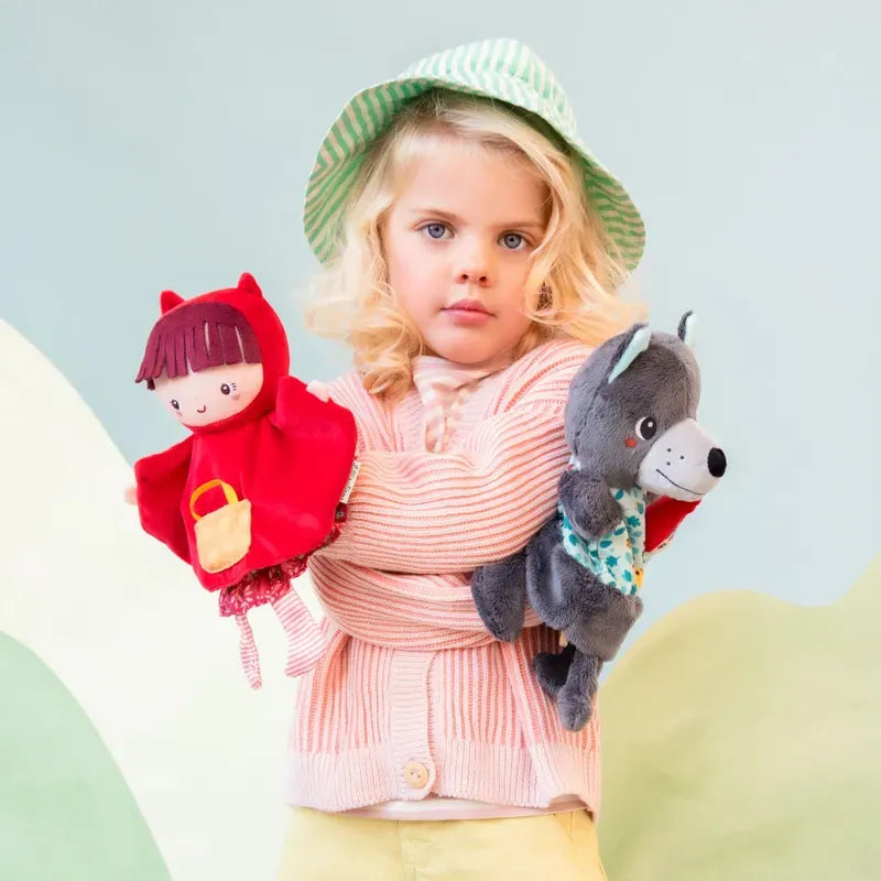 A young child with wavy blonde hair wearing a green-striped hat and a pink cardigan holds two puppets: a girl in a red outfit representing Lilliputiens Red Riding Hood Handpuppet and a gray puppet representing the Big Bad Wolf. The backdrop features pastel hills, perfect for imaginative and creative play.