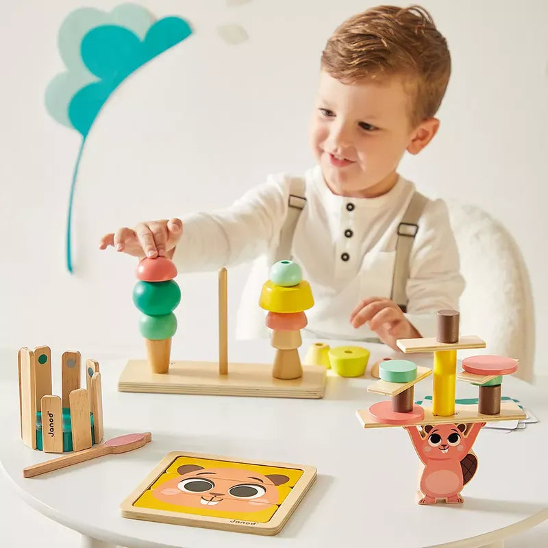 A young child with light brown hair plays enthusiastically on a white table. The child is engaging with colorful wooden games and educational toys, including the Janod Box of Toys 36 Months. The background features a simple, modern design with a wall accent resembling a tree.