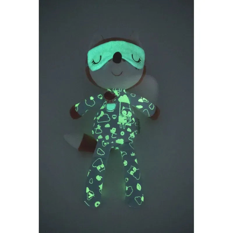 A Lilliputiens Alice Night Friend, resembling an Alice soft toy, is wearing glow-in-the-dark pyjamas with a sleep mask. The pajamas feature a pattern of clouds, stars, and other shapes. The toy is lying on a dark background, making the glowing patterns stand out. Plus, it's machine-washed for convenience.