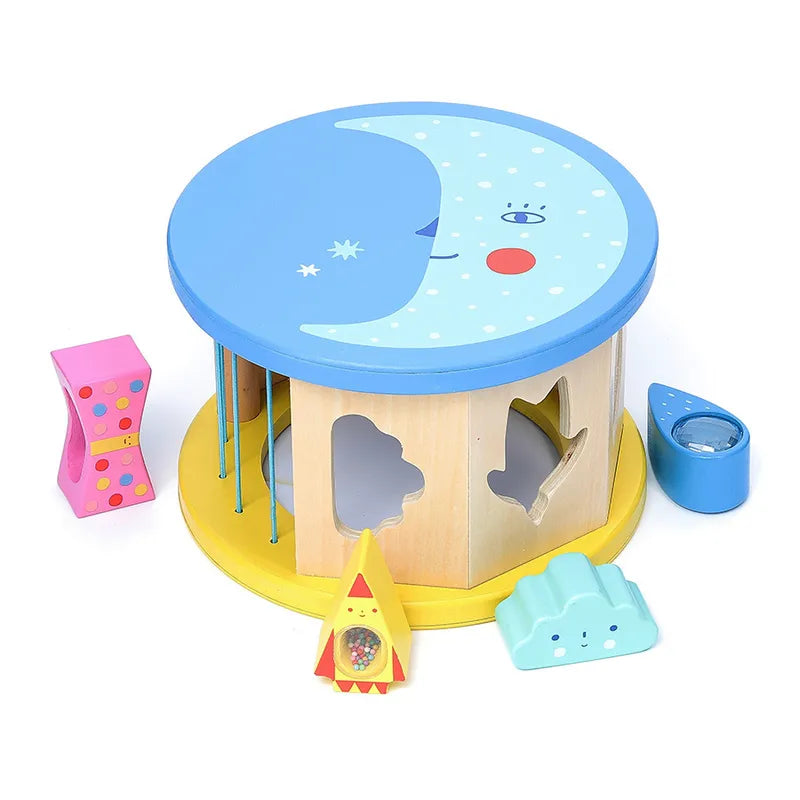 The Vilac Night and Day Shape Sorter is a vibrant wooden toy designed with a blue and yellow round base adorned with charming star and moon designs. Ideal for early learning, this set includes various geometric shapes like a rocket, cloud, hourglass, and teardrop all featuring cute faces. These shapes fit into corresponding cutouts on the base, making it perfect for enhancing fine motor skills.