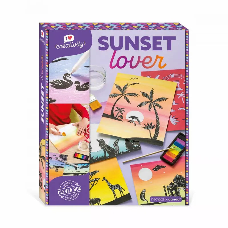 A colorful children's art kit box labeled "Janod Sunset Lover" showcases various sunset-themed art projects on the front. The artwork includes silhouettes of palm trees, giraffes, and flamingos against vibrant sunset backgrounds. The kit includes a watercolor paint set and brushes, perfect for sunset painting craft sessions.